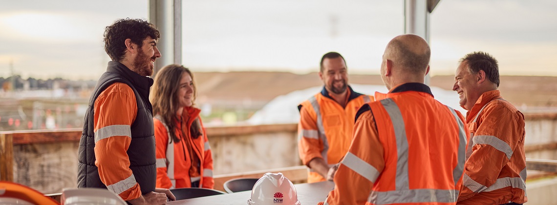 Five workers wearing high-vis orange vests talking around a table